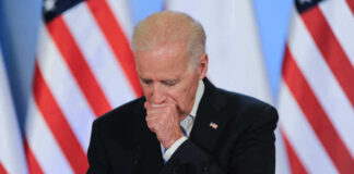 Joe Biden coughing in a suit standing in front of flags.