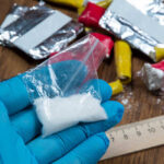 Shocking Discovery During DRUG RAID! Blue-gloved hand holding bag of white powder, other substances nearby.