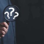 Hand holding magnifying glass with question marks inside.