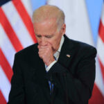 Joe Biden coughing in a suit standing in front of flags