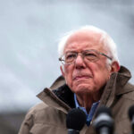 Bernie Sanders Attacks Free Market Innovation Bernie Sanders wearing glasses speaking outdoors with microphones.