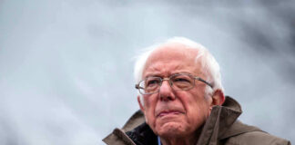 Bernie Sanders wearing glasses speaking outdoors with microphones.