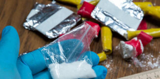 Blue-gloved hand holding bag of white powder, other substances nearby.