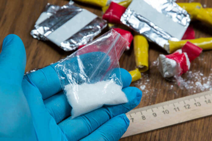 Blue-gloved hand holding bag of white powder, other substances nearby.