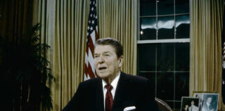 Ronald Reagan speaking at a desk holding a paper.