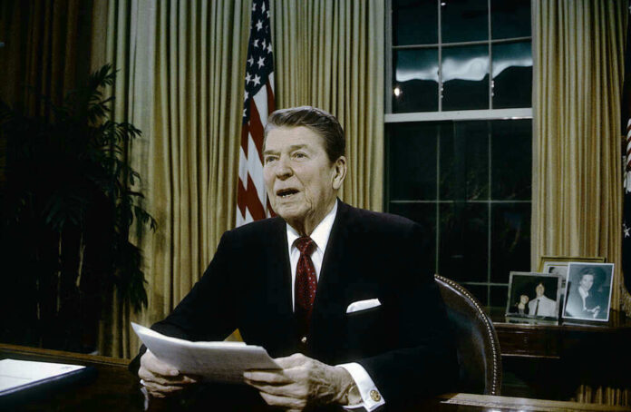 Ronald Reagan speaking at a desk holding a paper.