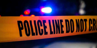 Police tape with flashing lights in the background.