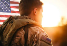 Soldier in camouflage facing American flag at sunset.