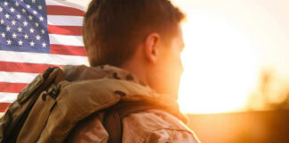 Soldier in camouflage facing American flag at sunset.