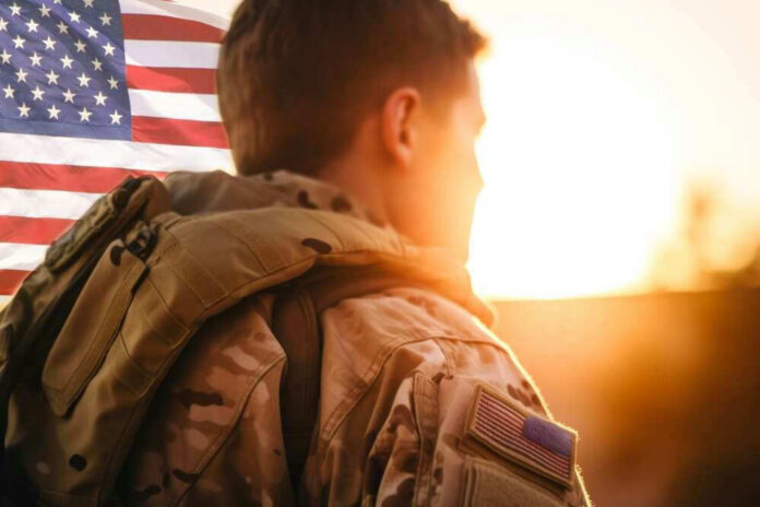 Soldier in camouflage facing American flag at sunset.