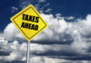 Yellow road sign reading taxes ahead clouds background
