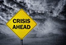 A yellow warning sign that reads 'CRISIS AHEAD' against a stormy sky