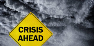 A yellow warning sign that reads 'CRISIS AHEAD' against a stormy sky