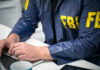 Person in FBI jacket working on laptop.