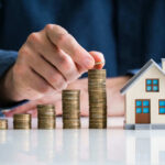Property Costs Trigger Nationwide Nightmare Hand stacking coins beside a miniature house model.