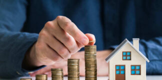 Hand stacking coins beside a miniature house model.