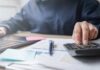 Water Bills EXPLODE — Families Can’t Afford Person using a calculator while working on a laptop and reviewing documents