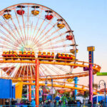 Upside-Down Launch At 100 MPH? Colorful amusement park scene featuring a ferris wheel and roller coaster at sunset