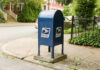 Stamp Shock Hits Sunday Blue USPS mailbox on a residential street corner.