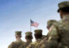 Trump Threat: Troops Out, Greenland In Soldiers in uniform facing American flag outdoors.