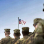 Trump Threat: Troops Out, Greenland In Soldiers in uniform facing American flag outdoors.