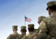Soldiers in uniform facing American flag outdoors.