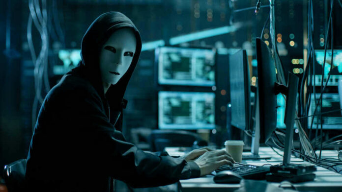 shutterstock_680078920 Person in hoodie and mask using computer, multiple screens.