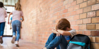 Sad child sitting against brick wall, kids running away.