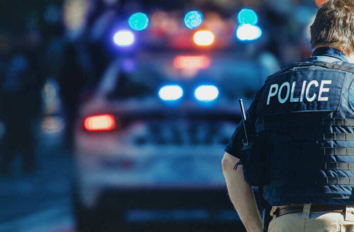 shutterstock_1543591088-1 Police officer stands near patrol car with lights flashing.
