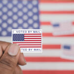 Barcode Ballots Ignite Mailroom Showdown Hand holding a vote-by-mail sticker with U.S. flag
