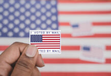 Hand holding a vote-by-mail sticker with U.S. flag