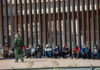 Surprise Deportation Pipeline Begins Border patrol officer watches group sitting by a fence.