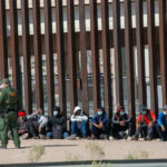 Surprise Deportation Pipeline Begins Border patrol officer watches group sitting by a fence.