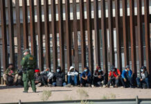 Surprise Deportation Pipeline Begins Border patrol officer watches group sitting by a fence.