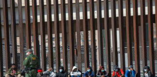 Border patrol officer watches group sitting by a fence.