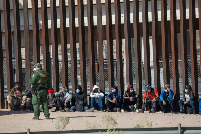 shutterstock_2079408463 Border patrol officer watches group sitting by a fence.