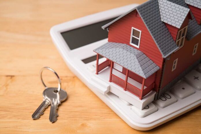 Red,House,Model,On,White,Calculator,And,House,Key,On A miniature red house on a calculator with keys nearby