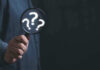 Hand holding magnifying glass with question marks.