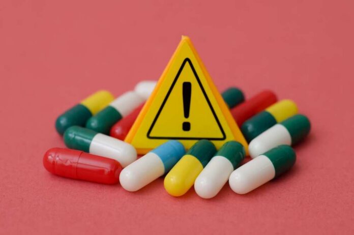 Yellow,Warning,Exclamation,Sign,Surrounded,By,Medical,Capsules,-,Drug Warning sign among assorted capsules on pink background
