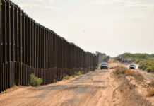Judge SLAMS Border Build – Why Now? A long border wall stretching through a desert landscape with vehicles nearby