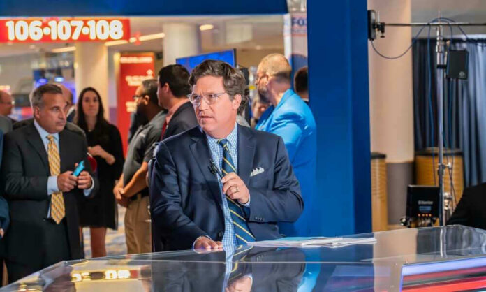 shutterstock_2329709309 Tucker Carlson at a broadcast desk with a microphone in a busy venue